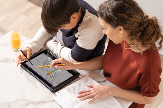 A male child works on a tablet while his female parent looks over his shoulder